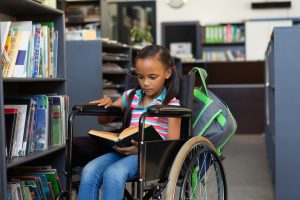 Front view of a disabled mixed-race schoolgirl reading a book in the library at school
