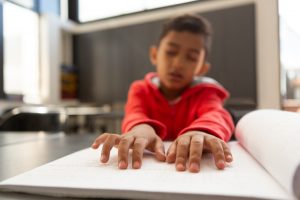 Schoolboy hands reading a braille book at desk in a classroom at elementary school